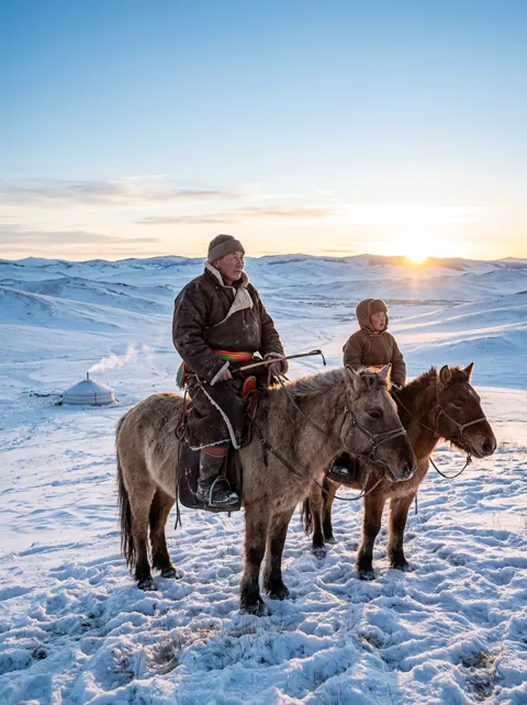 Father and son herders on winter horseback against vast Mongolian sunrise steppe